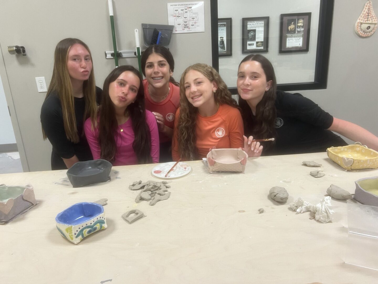 Five young women smiling together at a craft table with clay and tools.
