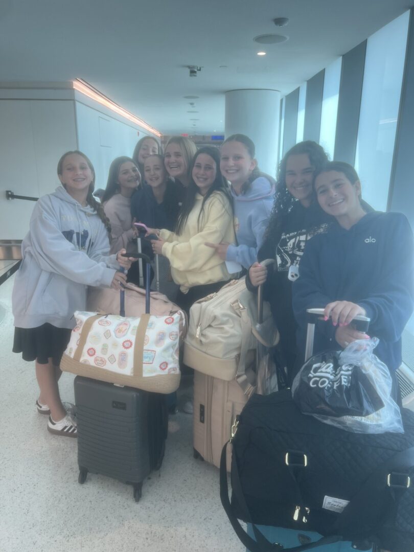 A group of seven people smiling with luggage in a bright room.