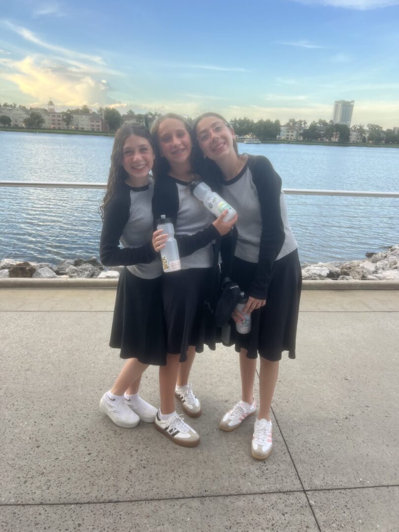 Three girls in black skirts and white shoes posing by a waterfront.