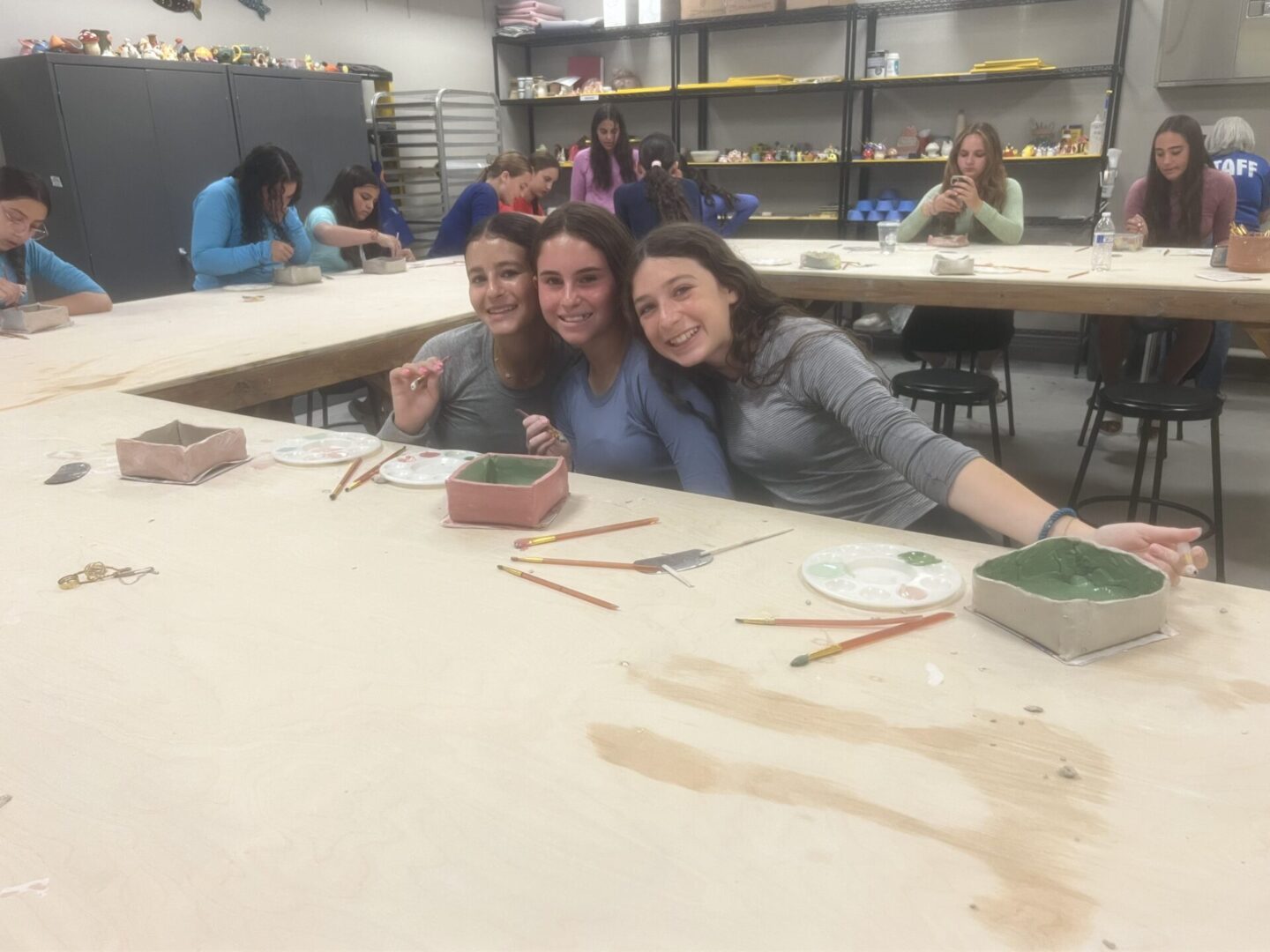 Three young women sitting at a pottery workshop table with clay and tools.