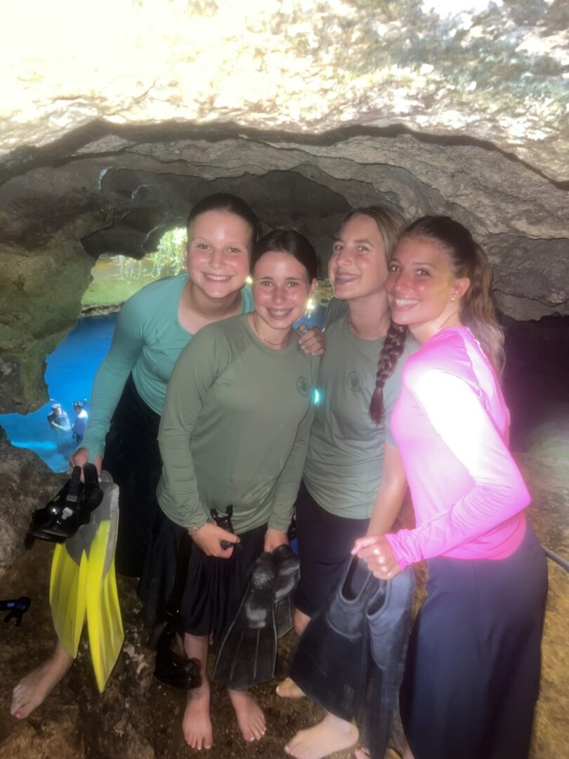 Four women smiling inside a cave, wearing casual outdoor clothing.