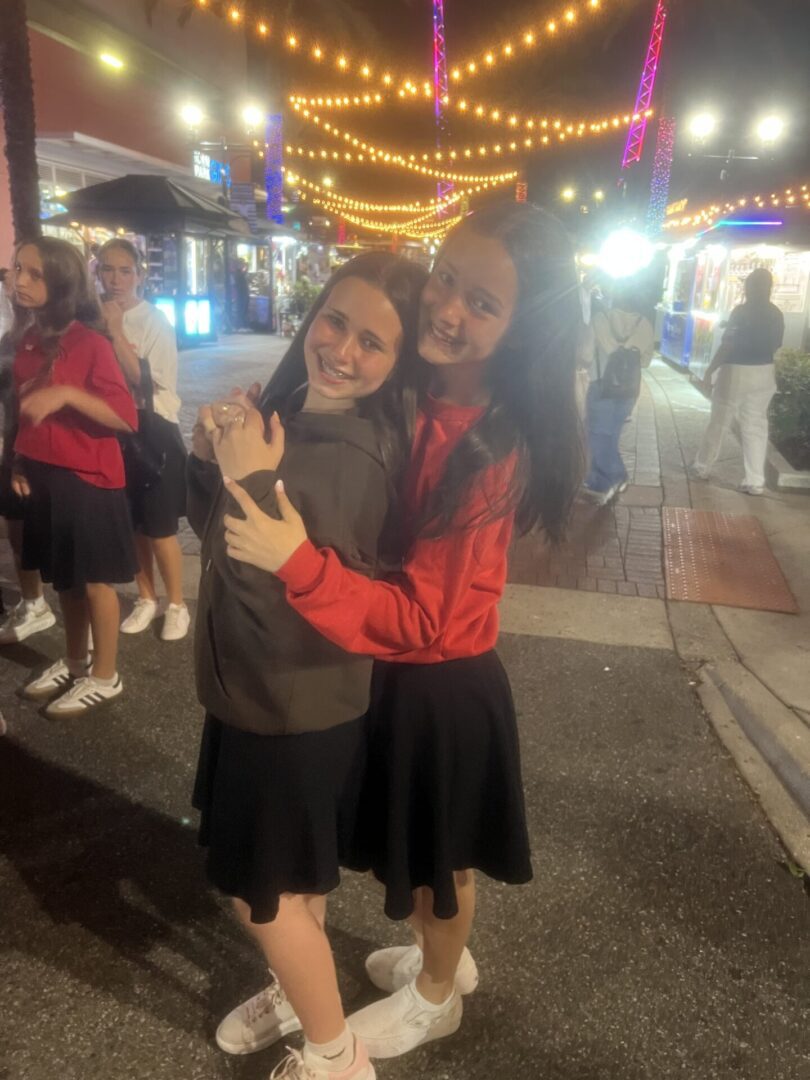 Two women hugging and smiling in a lively, lit street at night.