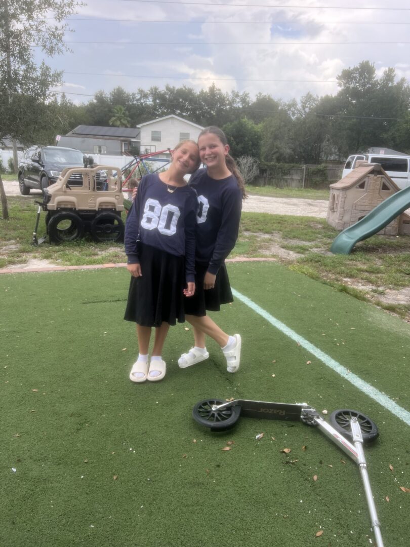 Two girls in matching sports jerseys posing on a field with lacrosse gear nearby.