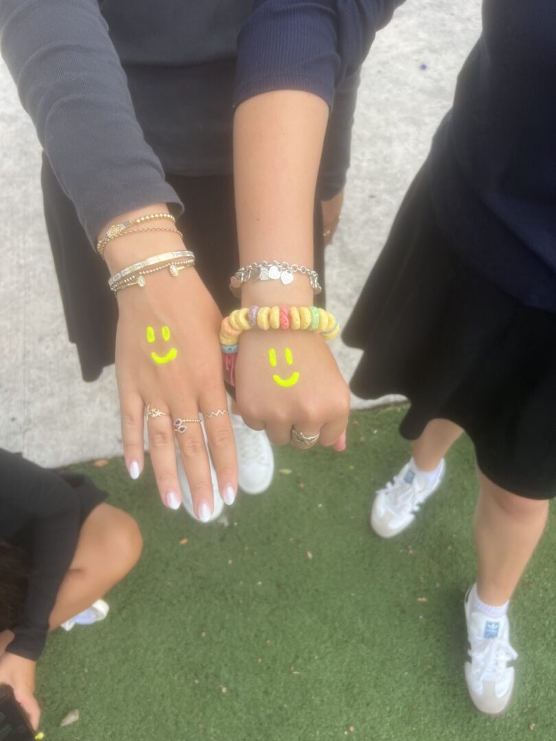 Two people show their hands with smiley face drawings and bracelets outdoors.