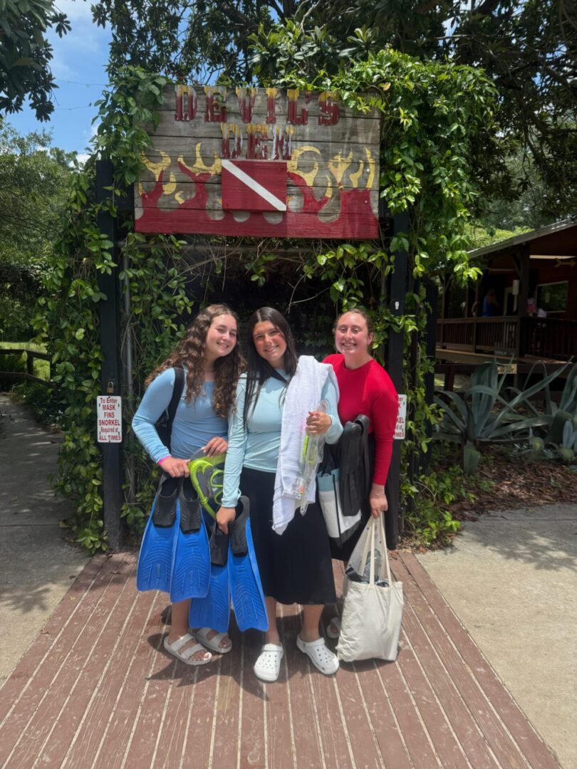 Three women posing happily outdoors with greenery and a sign in the background.