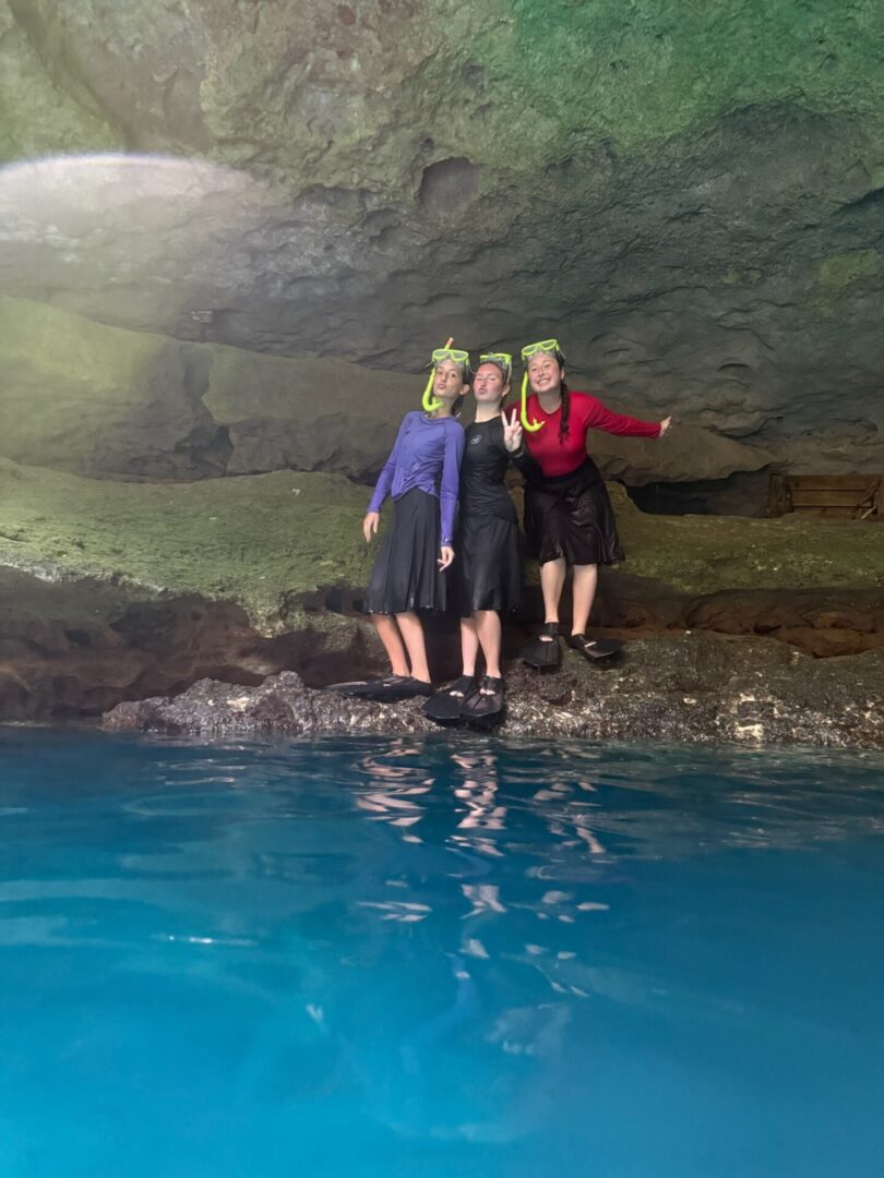 Three people standing on rocks inside a cave above clear blue water.