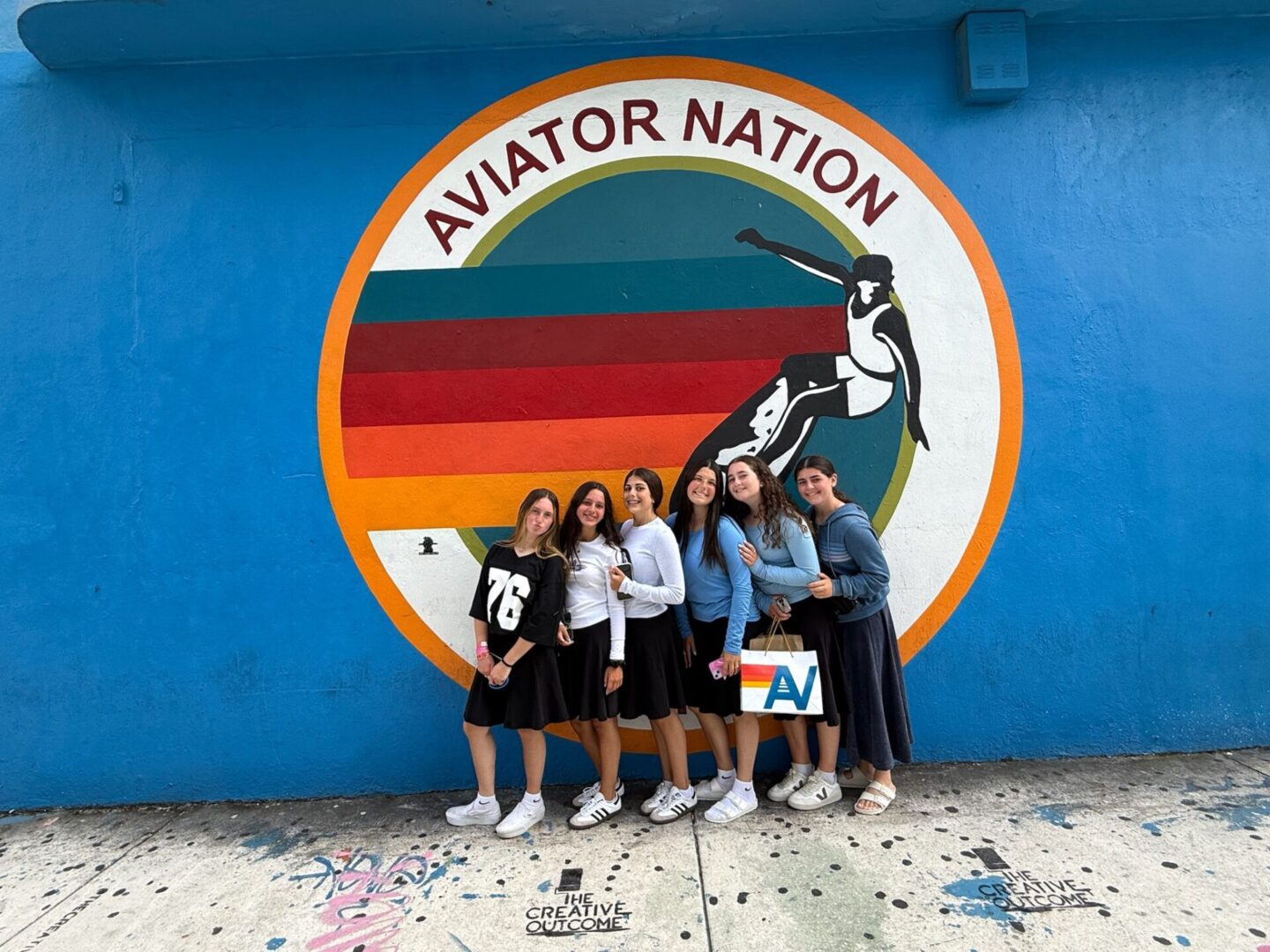 Group of six friends posing in front of an Aviator Nation mural.