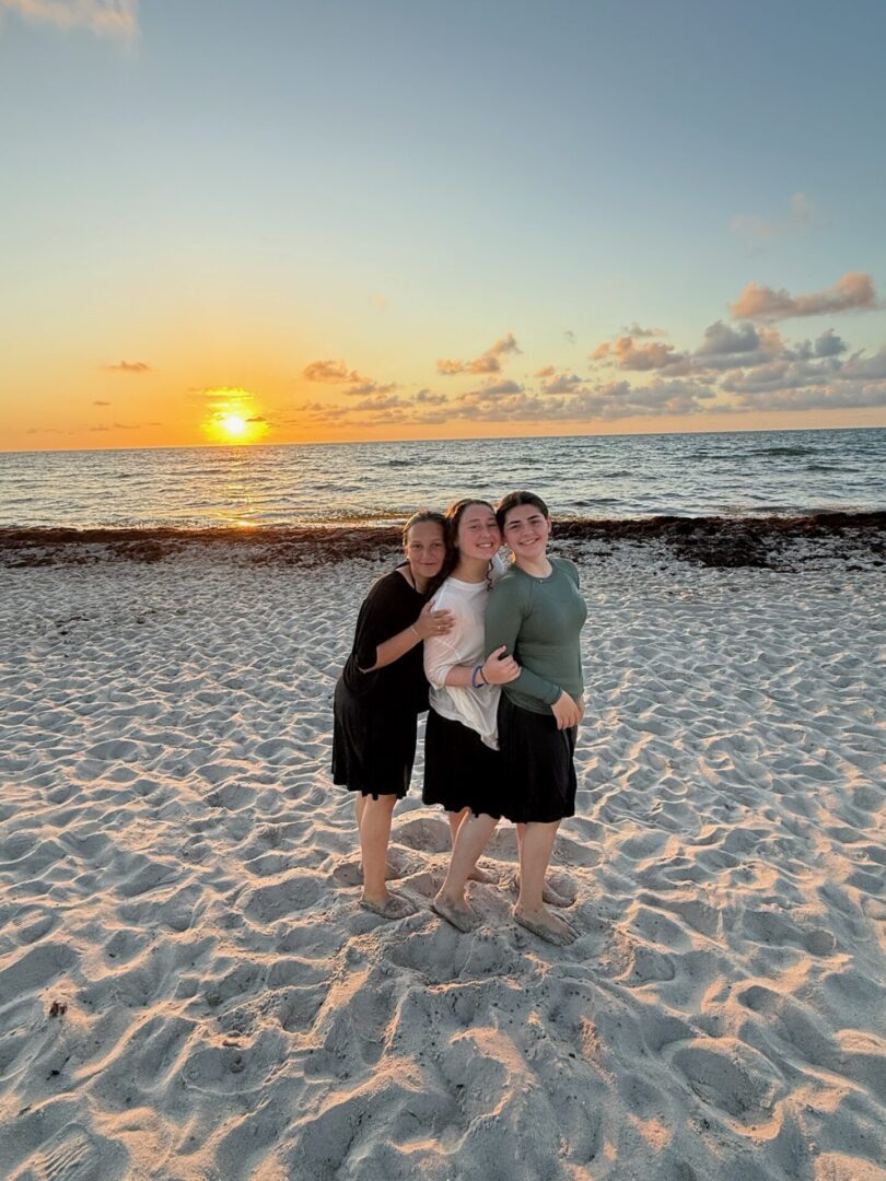 Three people smiling together on a beach at sunset.